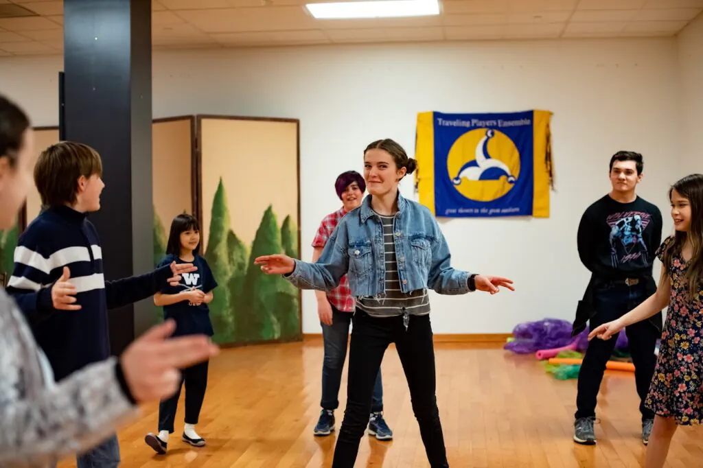 A group of students ages 8-18 stand in a circle playing an improv game. At the center is a 13 year old girl, with a playful smile.