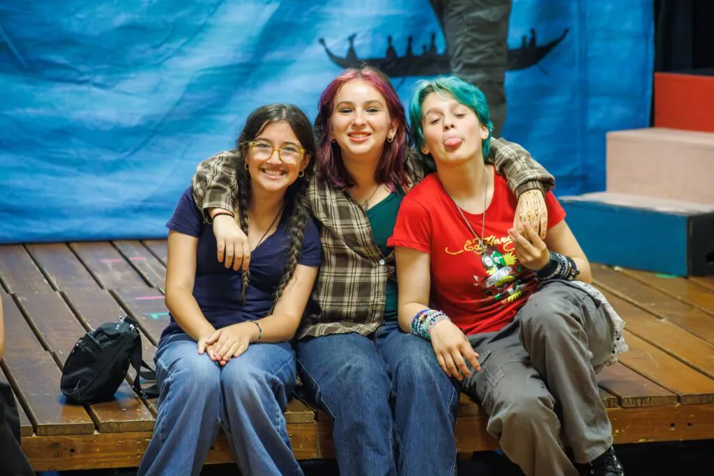A trio of 3 middle schoolers sit on the edge of the stage. They have their arms around each other and are smiling or making a goofy face at the camera.