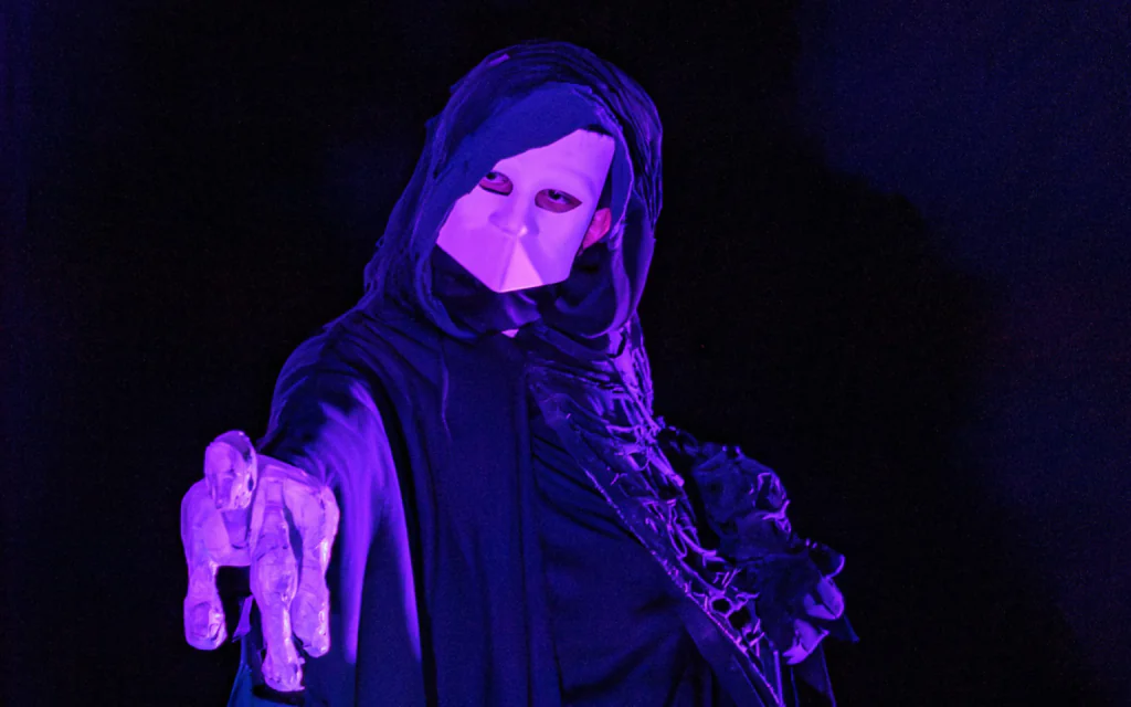 A performer dressed in a black hooded robe and a white bauta mask stares menacingly at the camera. They are holding a large skeletal hand made from papier mâché that is pointed toward the camera.