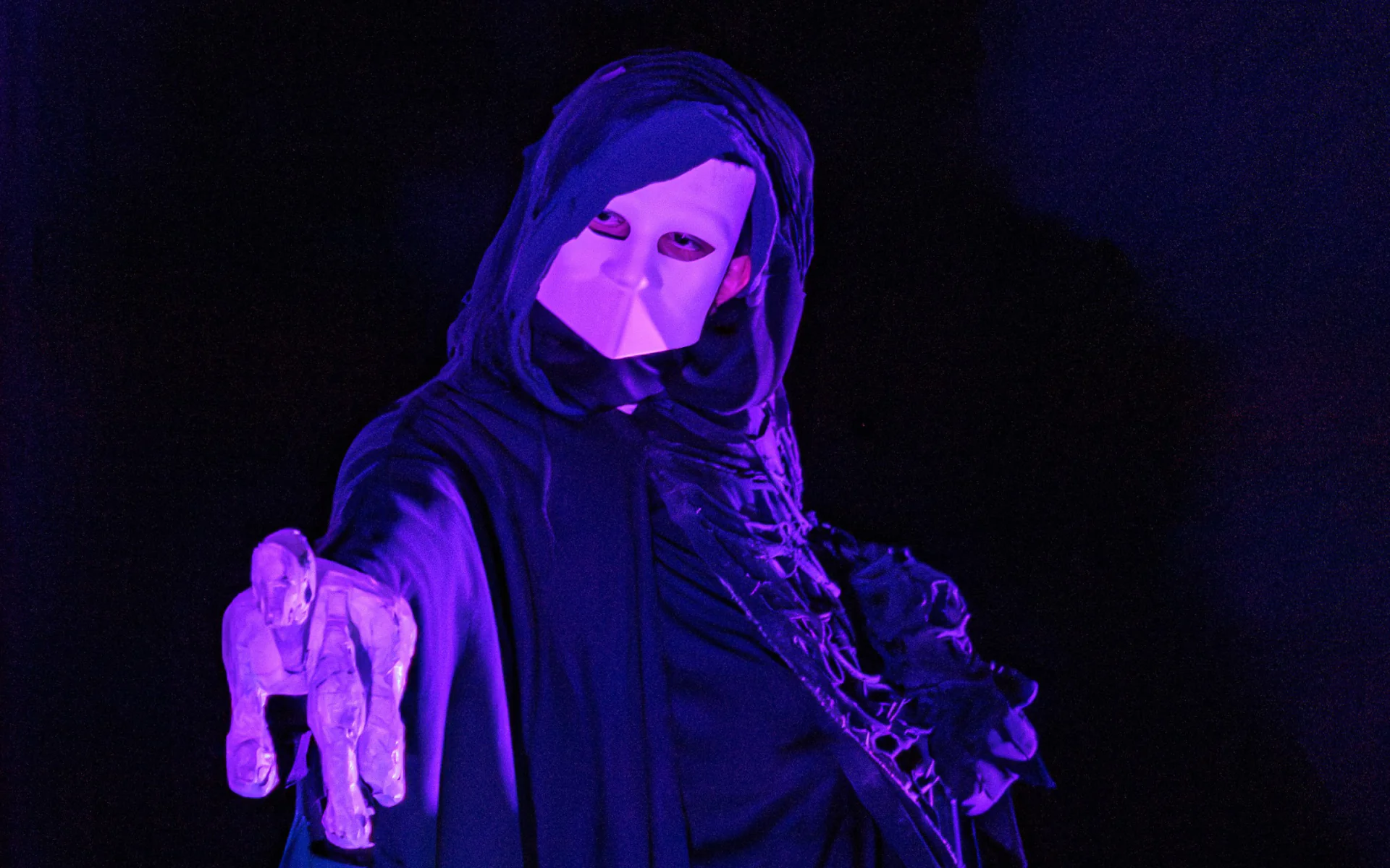 A performer dressed in a black hooded robe and a white bauta mask stares menacingly at the camera. They are holding a large skeletal hand made from papier mâché that is pointed toward the camera.