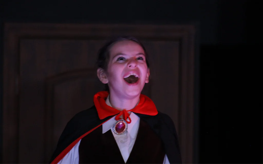 A middle school aged girl dressed as Count Dracula. The lighting is dark and spooky with a slight red cast to it. She is looking to the sky and laughing maniacally.