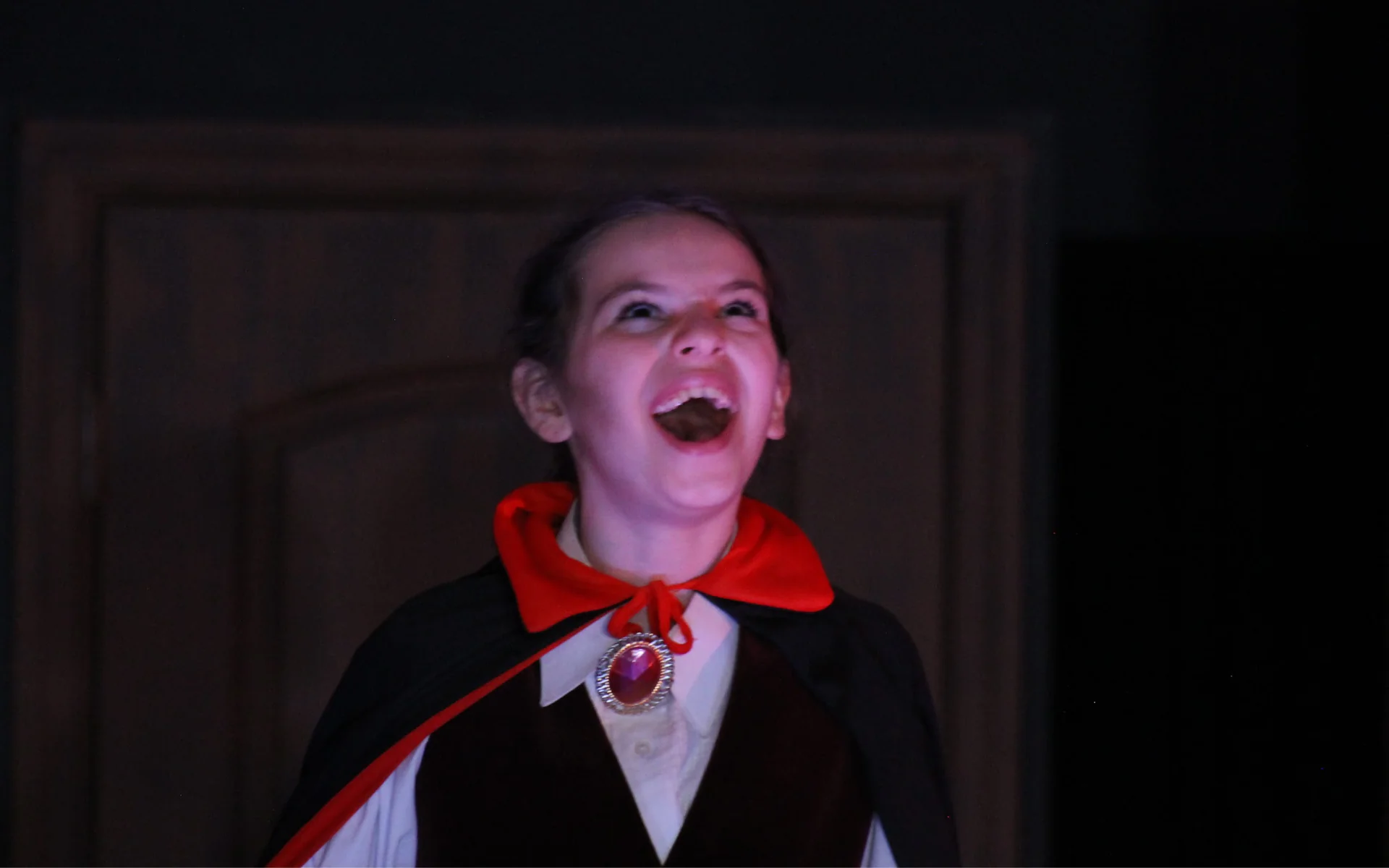 A middle school aged girl dressed as Count Dracula. The lighting is dark and spooky with a slight red cast to it. She is looking to the sky and laughing maniacally.