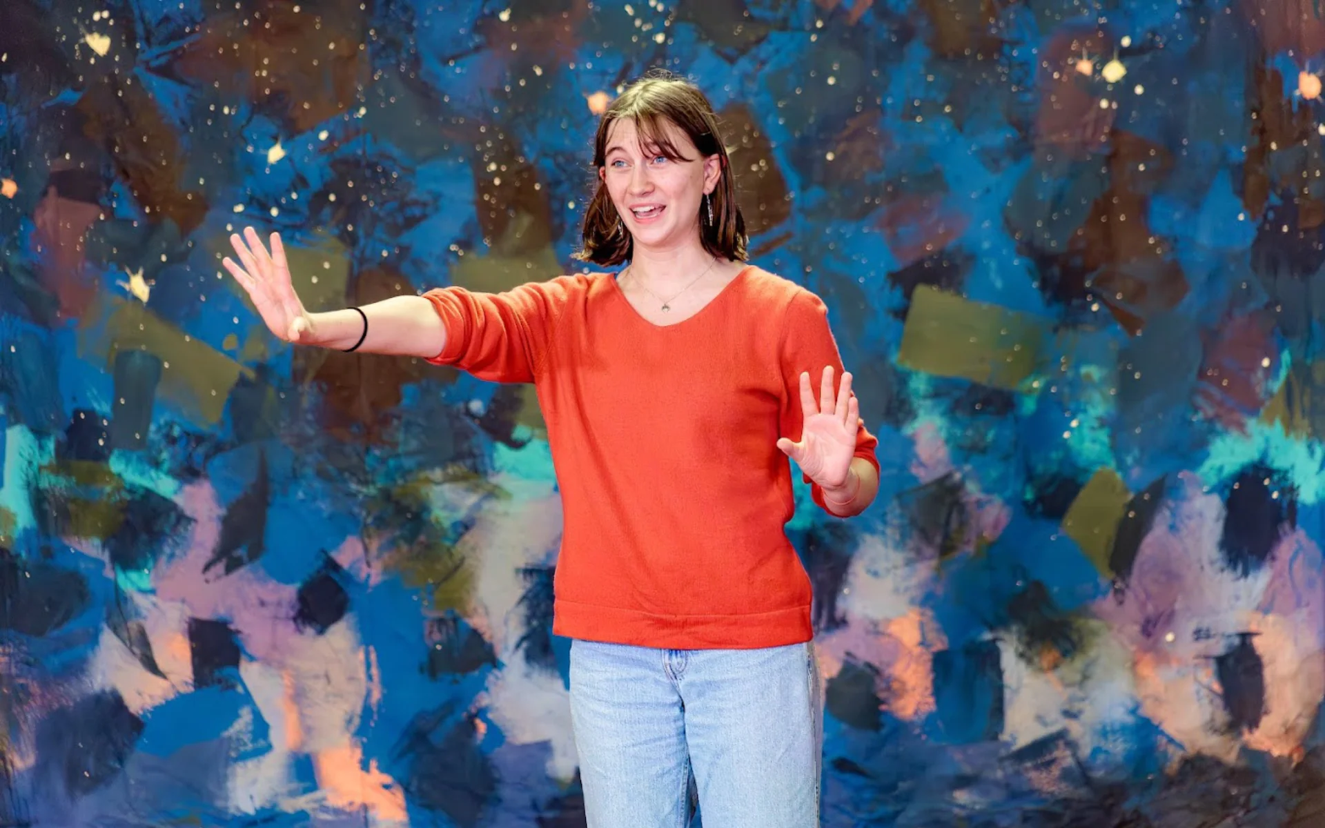 A high school aged girl stands center stage in front of a blue painted background. She is wearing a red sweater and gesturing excitedly as she performs a monologue.